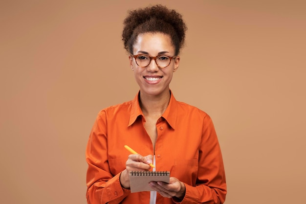 Cheerful woman wearing eyeglasses smiles at camera posing with notepad isolated beige background