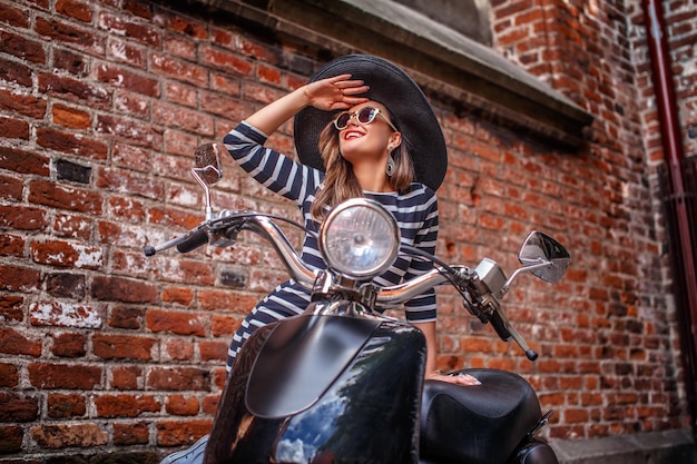 Donna allegra che indossa abiti di moda appoggiata su un classico scooter italiano nero e distoglie lo sguardo su una vecchia strada.