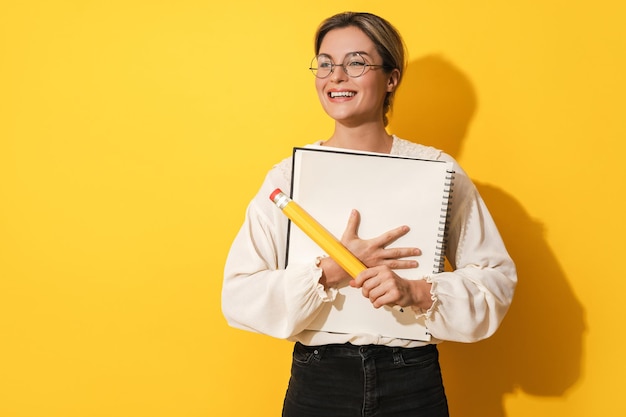 Cheerful woman wearing glasses holding big pencil and notebook on yellow background