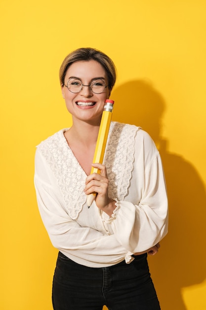 Cheerful woman wearing glasses holding big pencil on yellow background