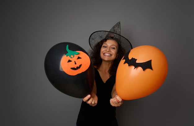 Cheerful woman wearing a wizard hat poses with black and orange air balloons with felt-cut bat and pumpkin, smiles toothy smile looking at camera. Halloween concept on grey background, copy space