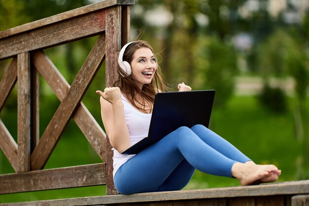 Cheerful woman in wireless headphones makes telework with laptop outdoors