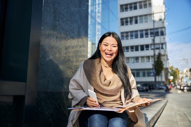 Cheerful woman with cellphone and papers sitting on steps outdoors