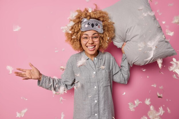 Photo cheerful woman with curly blode hair foolishes around has pillow fight after awakening wears comfortable pajama and blindfold surrounded by feathers floating in air isolated over pink background