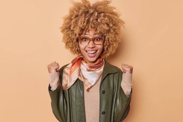 La donna allegra con i capelli ricci folti stringe i pugni celebra il successo sorrisi boadly mostra denti bianchi indossa occhiali giacca di pelle e fazzoletto intorno al collo isolato sopra la parete beige dello studio