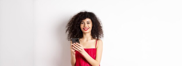 Photo cheerful woman with curly hair using smartphone in red dress smiling at camera standing over white b