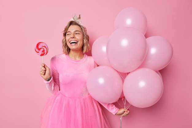 Cheerful woman with hairstyle wears small crown on head festive dress holds round caramel candy and bunch of inflated balloons enjoys celebration of special occasion poses against rosy background