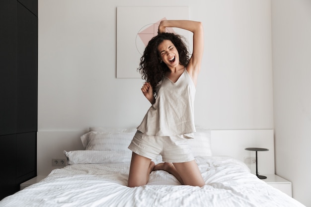 cheerful woman with long curly hair having fun and dancing, while sitting on bed with white clean linen in bedroom