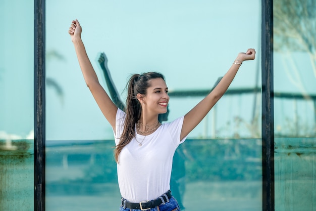 Cheerful woman with raised arms against glass wall