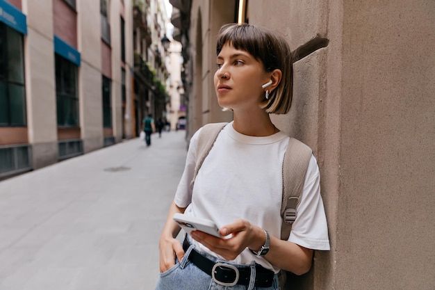 Donna allegra con smartphone in cuffie wireless sorridente in città signora con capelli corti in maglietta bianca jeans jeans in posa fuori sulla vecchia strada europea