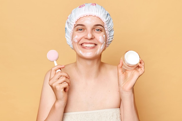 Cheerful woman woman wearing shower cap standing isolated over beige background holding facial cream and cleansing brush doing cosmetology procedures after taking shower