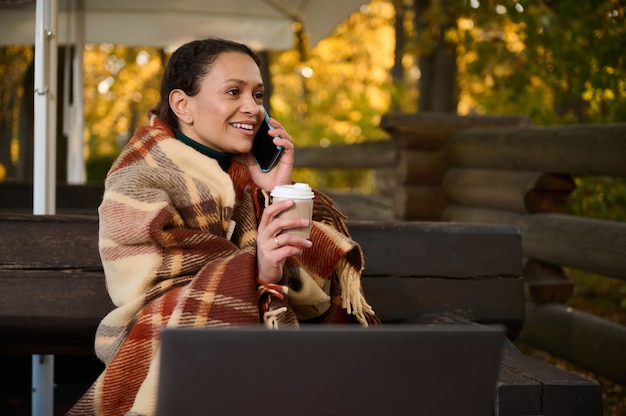 Cheerful woman wrapped in checkered woolen blanket, sitting on wooden bench in front of laptop screen monitor, holds a cardboard cup oh hot drink, talks on mobile phone in country house wooden terrace