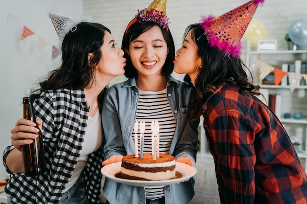 Cheerful women celebrating birthday while holding a birthday
cake with fire candles and kissed by girl best friends. young
ladies with funny party hats holding beer in decorated living room
at home.