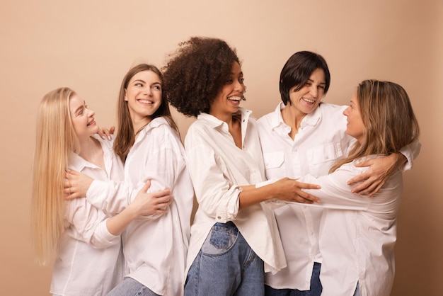 Cheerful women of different nations and ages in white shirts laugh looking at each other on brown background