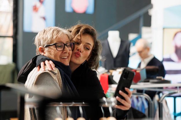 Cheerful women taking selfie with modern phone, smiling at camera in clothing store. Customers shopping for casual wear, buying stylish clothes and fashionable merchandise. Fashion concept