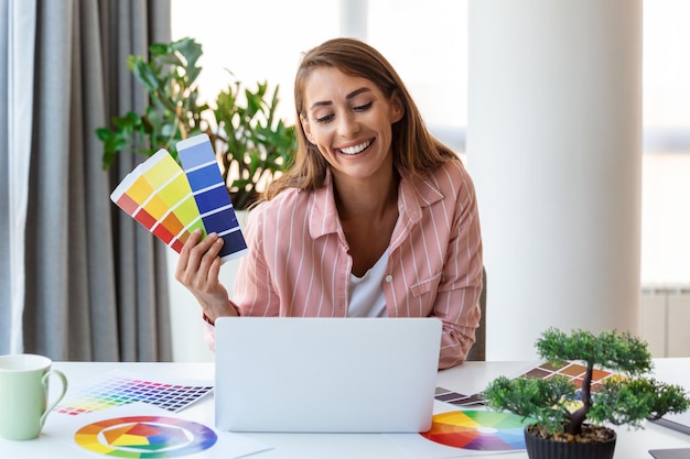 Cheerful youn woman designer having video conference with clients sitting at desk in front of computer holding color palettes gesturing and smiling copy space