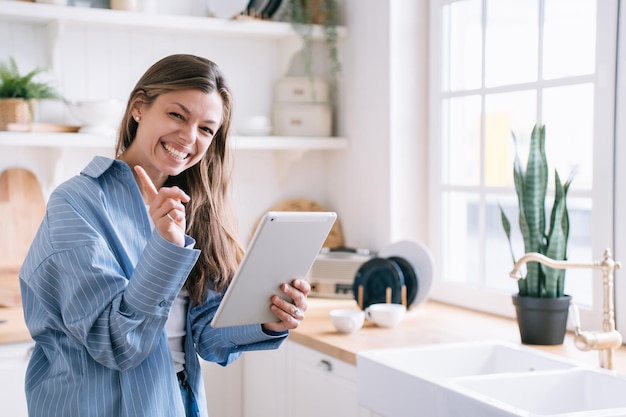 Cheerful young adult caucasian woman in blue shirt holds tablet\
toothy smiles looks at camera warns by index finger not to look at\
her messages tablet screen secret concept girl smiles at\
kitchen