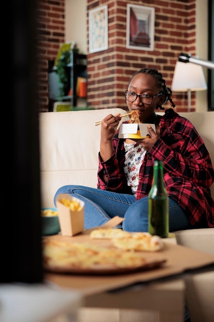 Cheerful young adult eating noodles from takeout delivery package and relaxing on couch with movie on tv. Using choppsticks to eat takeaway fast food meal and drinking beer from bottle.