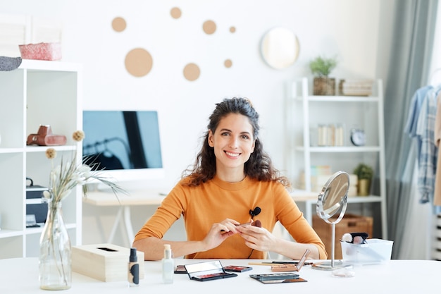 Cheerful young adult woman sitting at table with beauty products on it looking smiling