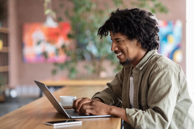 Cheerful young african american curly male typing on laptop at table with coffee cup and phone in