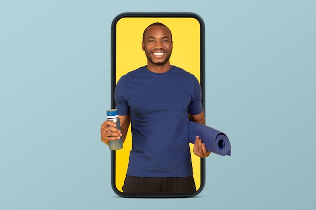 Cheerful young african american guy athlete coach in sportswear with mat and bottle of water on