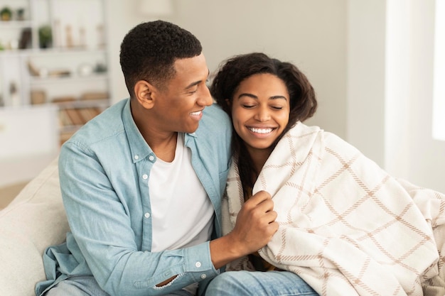 Cheerful young african american guy wraps lady in blanket couple enjoys warm together sitting on sofa