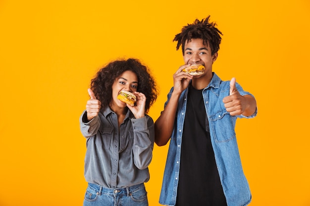 Cheerful young african couple standing isolated, eating burgers, thumbs up