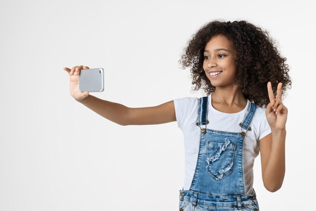 Cheerful young african girl kid using mobile phone taking selfie showing peace gesture