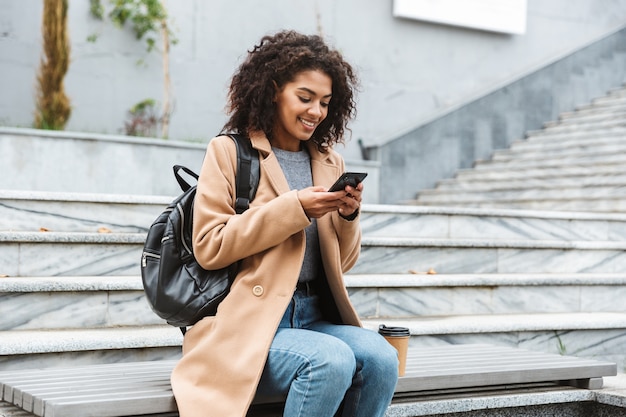 Cheerful young african woman wearing coat sitting outdoors, holding mobile phone