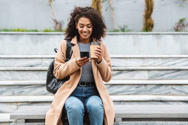 Cheerful young african woman wearing coat sitting outdoors, holding takeaway coffee cup