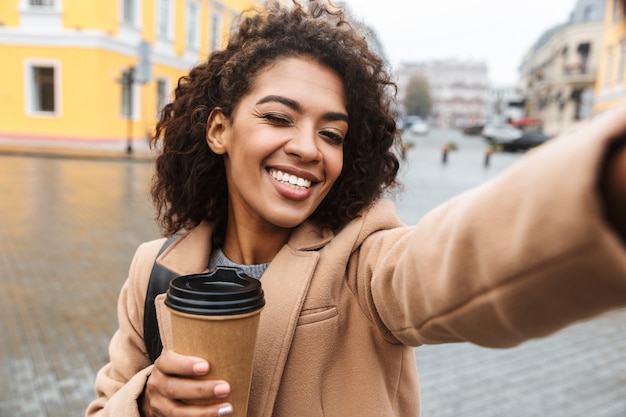 Photo cheerful young african woman wearing coat walking outdoors, holding takeaway coffee cup, taking a selfie