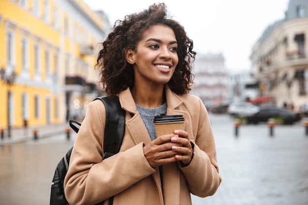 持ち帰りのコーヒーカップを持って、屋外を歩くコートを着て陽気な若いアフリカの女性