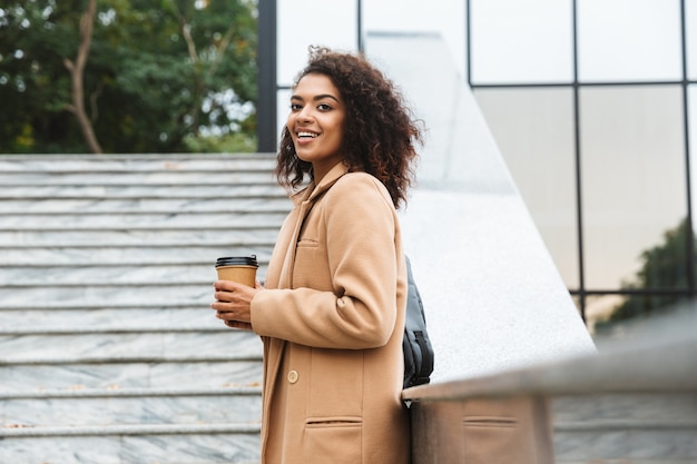 持ち帰りのコーヒーカップを持って、屋外を歩くコートを着て陽気な若いアフリカの女性