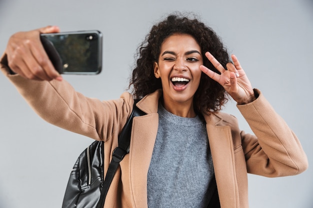 Cheerful young african woman wearing coat walking outdoors, taking a selfie