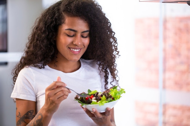 Foto allegra giovane donna afro-americana che mangia insalata di verdure nella cucina domestica - imagem.