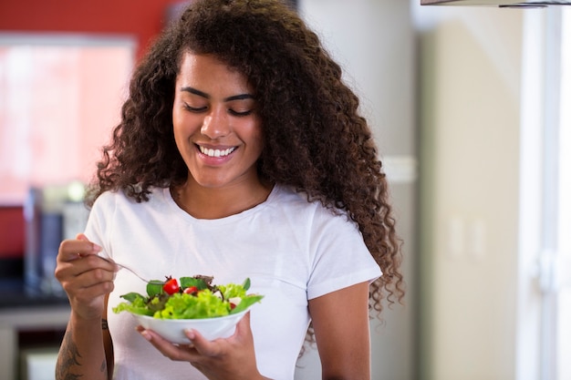 cheerful young afro american woman eating vegetable salad in home kitchen.