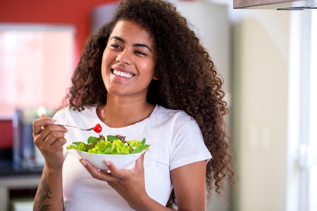 Allegra giovane donna afro americana che mangia insalata di verdure nella cucina di casa