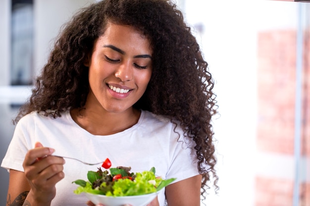 家庭の台所で野菜サラダを食べる陽気な若いアフリカ系アメリカ人女性