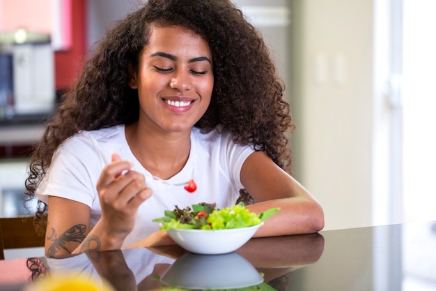 Photo cheerful young afro american woman eating vegetable salad in home kitchen