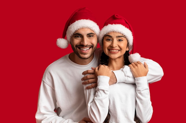 Cheerful Young Arab Couple In Santa Hats Embracing And Smiling At Camera