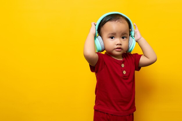 陽気な若いアジアの少年2歳の赤いTシャツの男の子黄色の背景に分離されたヘッドフォンでお気に入りの音楽を聴く母の日愛家族のライフスタイルのコンセプト