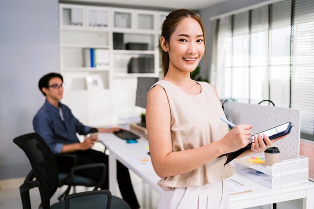 Photo cheerful young asian businesswoman entrepreneur in casual smiling and looking at the camera while holding a digital tablet and pencil happy businesswoman standing in the office of a modern workplace