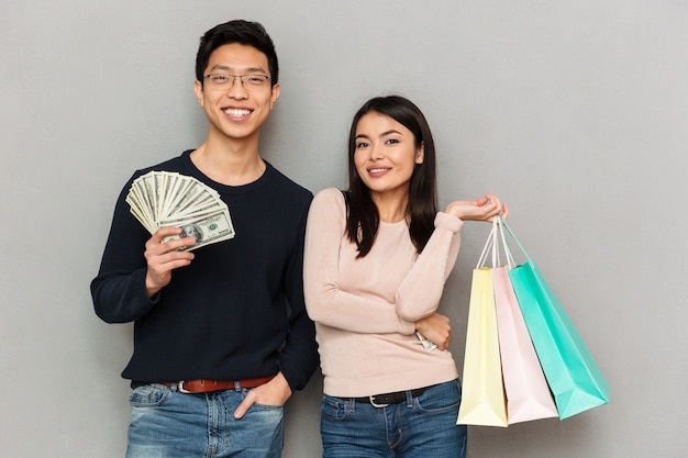 Cheerful young asian loving couple holding money and shopping bags.