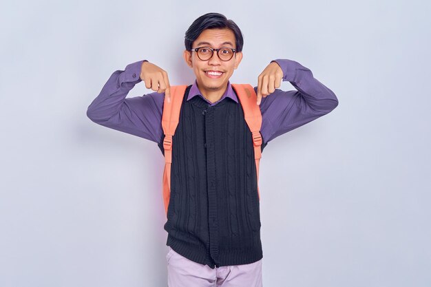 Cheerful young asian male student from college in casual\
clothes with backpack pointing at something with finger isolated on\
grey background education in high school university college\
concept