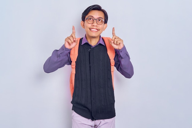 Photo cheerful young asian male student from college in casual clothes with backpack pointing at something with finger isolated on grey background education in high school university college concept