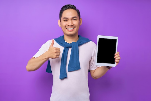 Cheerful young Asian man in casual clothes showing digital tablet with empty screen for mockup and thumb up gesture isolated over purple background