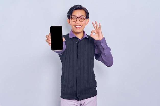 Cheerful young Asian man in casual shirt and vest showing mobile phone blank screen and making okay sign gesture isolated on white background People lifestyle concept