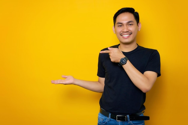 Cheerful young Asian man in casual tshirt pointing finger to his open hand palm isolated on yellow background Great offer concept