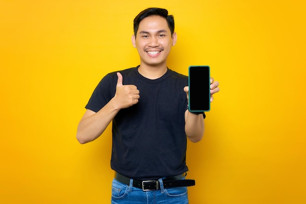 Cheerful young Asian man in casual tshirt showing mobile phone with blank screen showing thumb up gesture isolated on yellow background People lifestyle concept