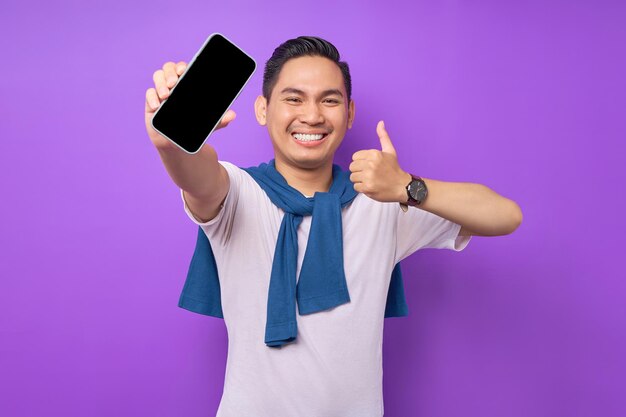 Cheerful young Asian man demonstrating smartphone with empty screen showing thumb up gesture isolated on purple background Advertising new mobile app mockup concept